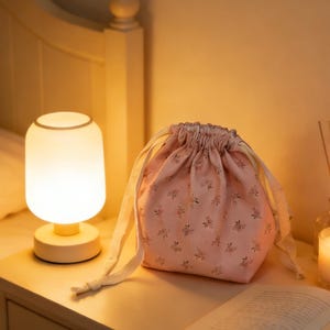 Pode incluir: Uma bolsa rosa com cordão e padrão floral está em uma mesa de cabeceira ao lado de uma pequena lâmpada e um livro aberto. A bolsa tem cordões brancos e uma parte superior franzida. A lâmpada tem uma cúpula branca e uma base creme.
