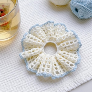May include: A handmade, off-white crochet scrunchie with a delicate, openwork design and a light blue trim. The scrunchie is circular, with a hole in the center, and sits on a white textured surface. Two balls of yarn are in the background.