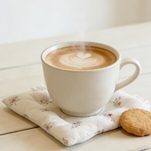 May include: A white ceramic mug filled with a latte, featuring a heart-shaped design in the foam, sits on a floral-patterned coaster. A golden-brown cookie rests beside the mug, creating a cozy, inviting scene.
