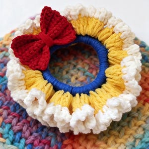 May include: A handmade crochet hair tie featuring a red bow, yellow, white, and blue accents. The hair accessory is set against a colorful, rainbow-hued crochet background. This item is perfect for adding a touch of handmade charm to any hairstyle.