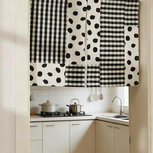 May include: Black and white patterned kitchen curtain. The curtain features a patchwork design with gingham and polka dot patterns. The curtain hangs in a kitchen doorway, with a glimpse of the kitchen interior visible in the background.