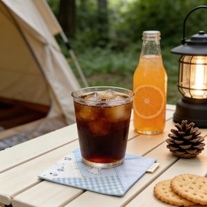 Puede incluir: Escena de camping con un vaso de cola con hielo sobre un posavasos azul y blanco. Una botella de refresco de naranja, una linterna encendida, un pi&ntilde;a y galletas est&aacute;n sobre una mesa de madera. Una tienda de campa&ntilde;a es visible en el fondo.