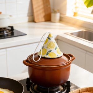 Op de afbeelding: Een bruine keramische pot met een deksel, bedekt met een pannenlap met citroenpatroon. De pot staat op een kookplaat met een brander aan. Een koekenpan met een gebakken ei en een broodmand zijn ook zichtbaar.