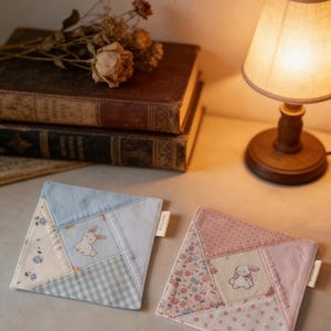 Op de afbeelding: Twee vierkante, gewatteerde onderzetters met een patchwork ontwerp. Eén is lichtblauw met een konijnengrafiek, de andere is roze met een konijnengrafiek. De onderzetters liggen op een lichtgekleurd oppervlak.