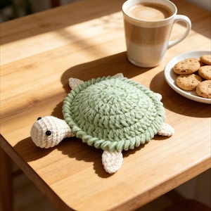 May include: A handmade crochet turtle coaster in light green and cream colors. The turtle has a rounded shell, four small feet, and a head with black eyes. The coaster is on a wooden table next to a mug of coffee and a plate of cookies.