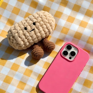 Può includere: Un giocattolo di peluche a forma di arachide beige all'uncinetto con occhi neri e un sorriso, e piedi all'uncinetto marroni. Una custodia per telefono rosa è accanto al giocattolo su una coperta da picnic a scacchi gialli e bianchi.