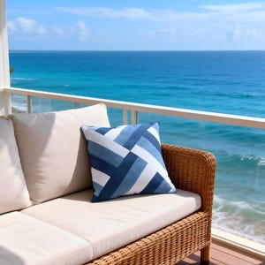 Op de afbeelding: Een rotan bank met cr&egrave;mekleurige kussens en een blauw en wit geometrisch patroon kussen staat op een houten dek met uitzicht op de zee. Het kussen heeft een ontwerp van diagonale strepen in blauwtinten. De achtergrond toont een helderblauwe lucht en zee.