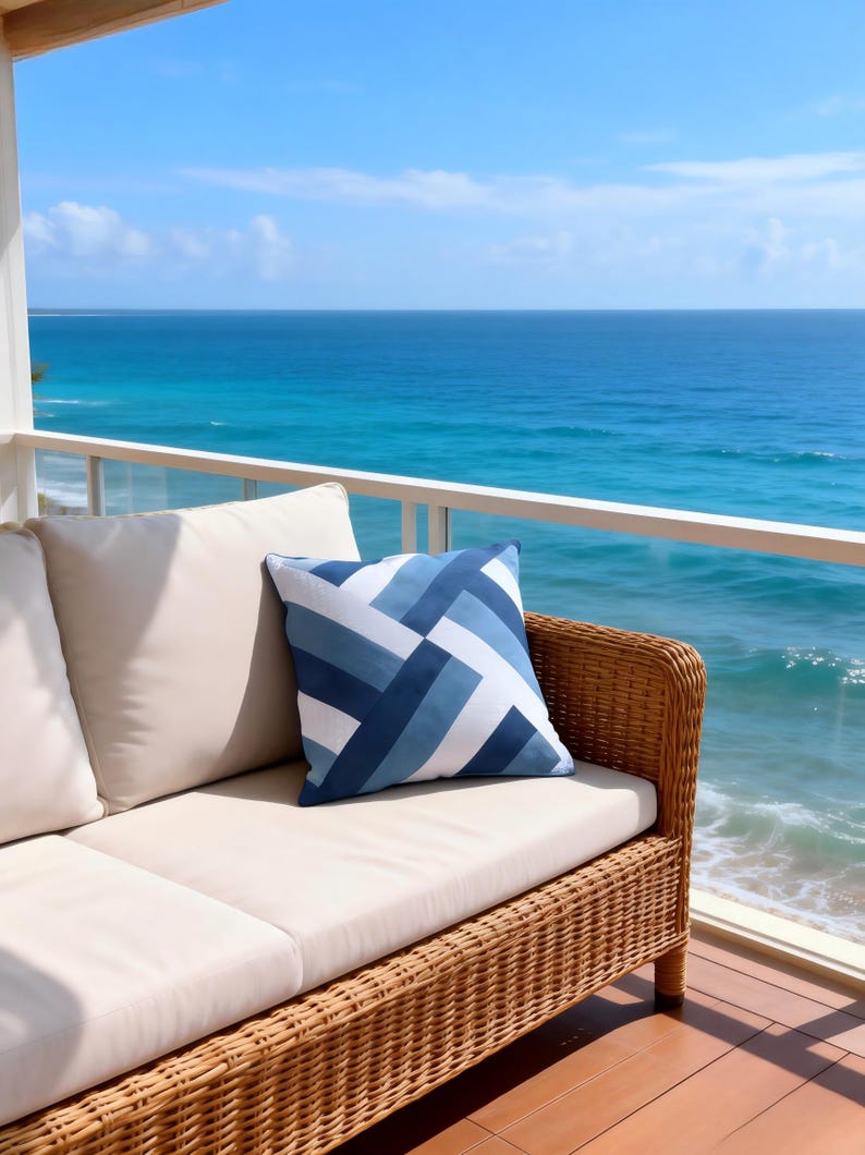 Op de afbeelding: Een rotan bank met cr&egrave;mekleurige kussens en een blauw en wit geometrisch patroon kussen staat op een houten dek met uitzicht op de zee. Het kussen heeft een ontwerp van diagonale strepen in blauwtinten. De achtergrond toont een helderblauwe lucht en zee.