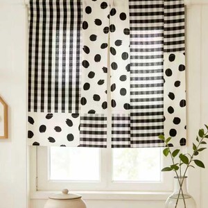 May include: Black and white patterned window curtains. The curtains feature a mix of gingham, polka dot, and striped patterns. The window is framed by a white trim. A ceramic jar with a lid and a wooden tray sit on a table in front of the window.