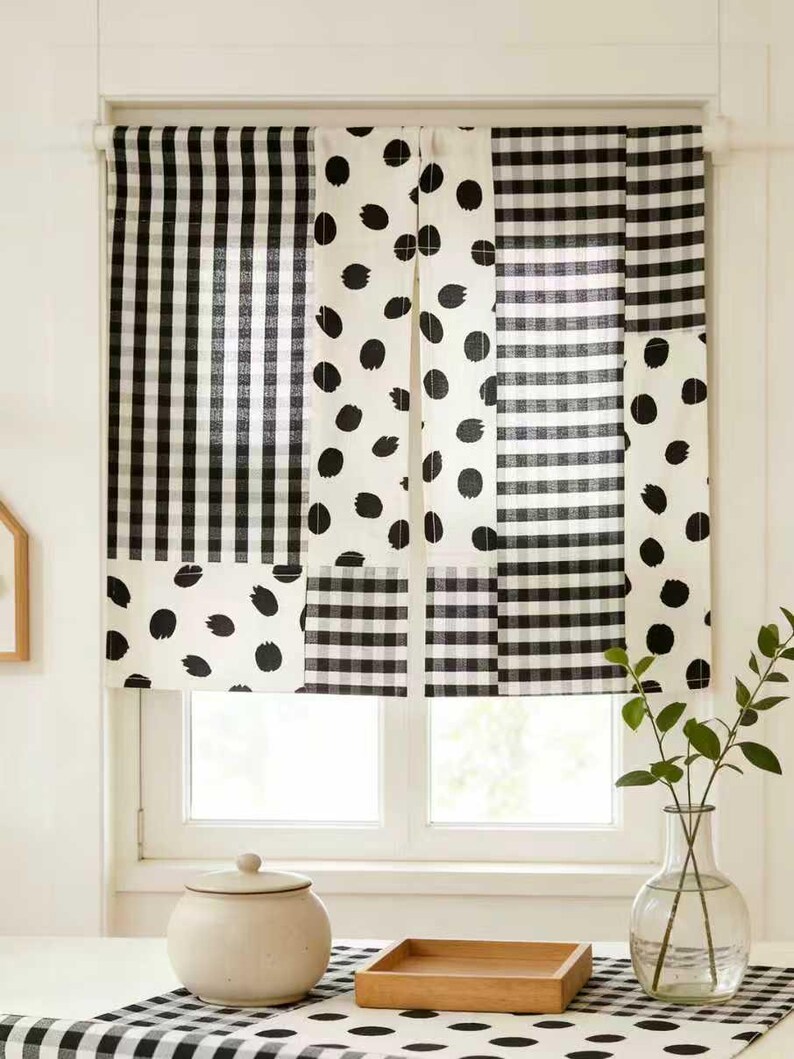 May include: Black and white patterned window curtains. The curtains feature a mix of gingham, polka dot, and striped patterns. The window is framed by a white trim. A ceramic jar with a lid and a wooden tray sit on a table in front of the window.