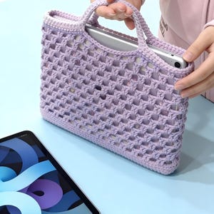 May include: A lavender crochet tablet bag with a handle, holding a silver tablet. The bag has a woven design and is held by a person. Another tablet is on a light blue surface.