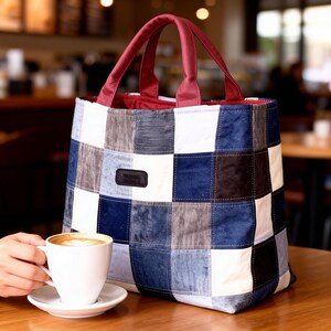 Op de afbeelding: Een patchwork tote bag met een geblokt patroon in tinten blauw, wit, grijs en zwart. De tas heeft bordeauxrode handvatten en een bordeauxrode binnenkant. Een witte koffiekop en schotel staan op een houten tafel.