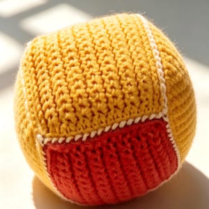 May include: A spherical ball crafted from knitted yarn, featuring a textured mustard yellow surface accented by a prominent square patch in a vibrant red hue. The edges of the red patch are defined by thick, white stitched detailing.