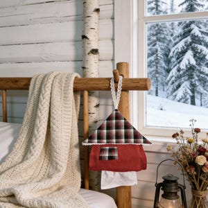 May include: A decorative house-shaped fabric item with a red body and a plaid roof hangs from a wooden bed frame. A white lace trim borders the roof. A cream-colored blanket and a vintage lantern are also visible.