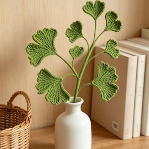 Könnte beinhalten: Ein gehäkelter Ginkgo-Blattzweig mit leuchtend grünen, fächerförmigen Blättern an einem zentralen Stiel. Die Textilpflanze steht aufrecht in einer glatten, matten weißen Keramikvase mit einem abgerundeten Boden und einem schmalen Hals.