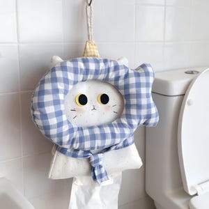 May include: A whimsical white cat-shaped tissue holder with a blue and white gingham patterned collar and headpiece. The tissue holder is hanging on a hook in a bathroom setting. The cat has black eyes and pink cheeks.