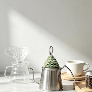 May include: A stainless steel gooseneck kettle topped with a sage green, cone-shaped crocheted lid cover featuring a small grey loop handle. The kettle sits alongside a clear glass coffee dripper, a glass carafe, a white ceramic mug, and a small jar.