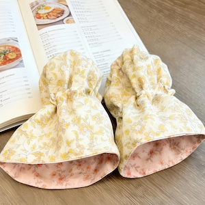 May include: Two floral fabric drawstring bags. The exterior fabric is cream-colored with a yellow floral pattern. The interior lining is a pink floral pattern. The bags are sitting on top of an open book.