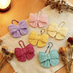 May include: A collection of six handmade crochet bow keychains in various pastel colors: purple, pink, yellow, red, and teal. Each bow has a beaded chain for attachment. The keychains are arranged on a neutral-colored cloth.