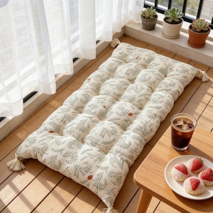 Könnte beinhalten: Ein rechteckiges, gestepptes Bodenkissen mit botanischem Muster in Creme und Grün, an jeder Ecke mit Quasten verziert. Das Kissen liegt auf einem Holzboden, neben einem kleinen Holztisch mit einem Teller Mochi und einem Glas Eiskaffee.