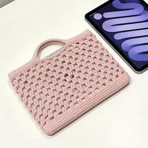 以下が含まれることがあります： 持ち手が付いた、薄いピンク色のオープン織りのかぎ針編みタブレットスリーブ。スリーブは長方形で、格子状のパターンと無地のボーダーがあります。黒いベゼルと紫色の画面を備えたタブレットがスリーブの隣にあります。