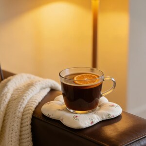 May include: A clear glass mug filled with a dark beverage and a slice of orange, resting on a floral-patterned coaster. A white knitted blanket with fringe is draped nearby. The scene is illuminated by a warm-toned lamp.