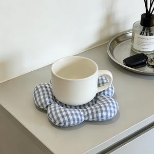 May include: A white ceramic mug rests on a blue and white gingham flower-shaped coaster. The coaster is made of soft fabric. The mug is on a gray surface, with a silver tray and diffuser in the background.