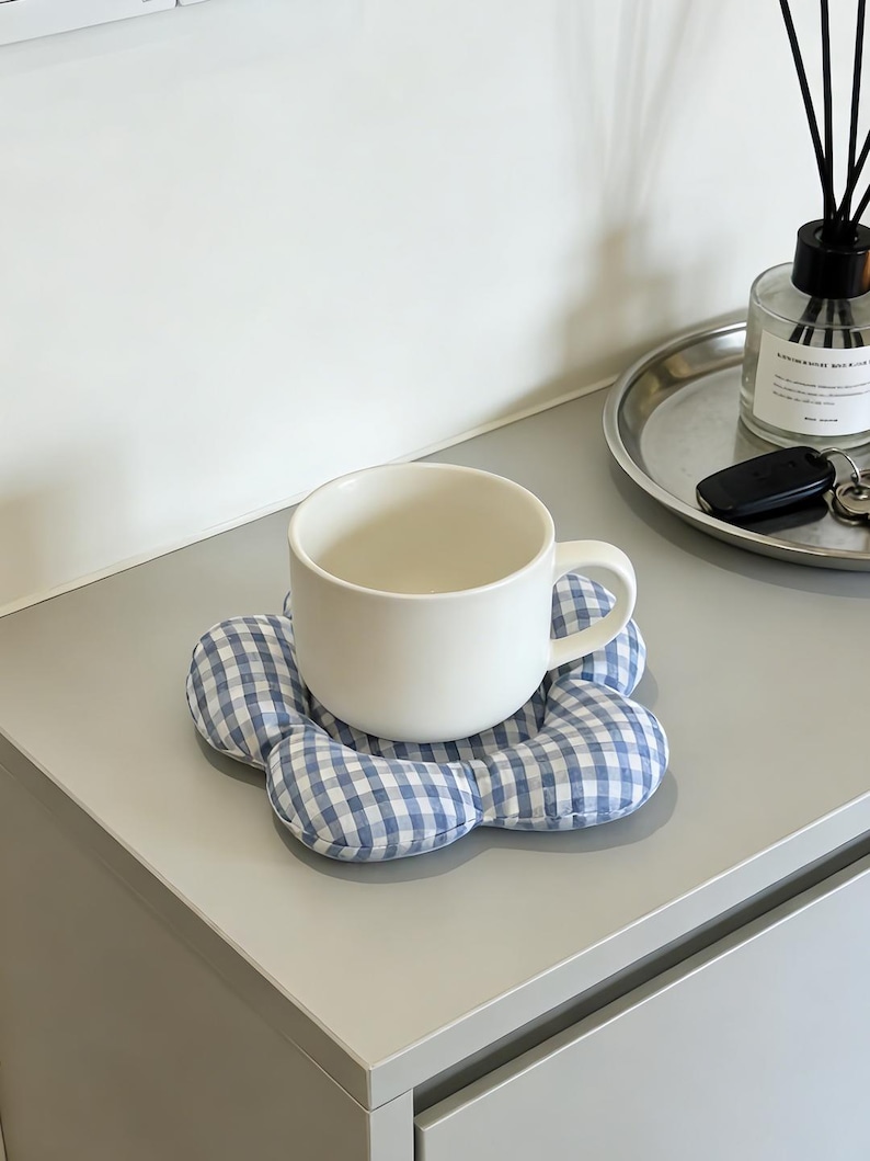 May include: A white ceramic mug rests on a blue and white gingham flower-shaped coaster. The coaster is made of soft fabric. The mug is on a gray surface, with a silver tray and diffuser in the background.