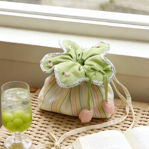 Può includere: Una borsa con coulisse con una parte superiore floreale verde e un corpo a righe. La borsa ha accenti rosa a forma di tulipano e un nastro color crema. Un bicchiere di palline di melone verdi e un libro sono anche nell'immagine.