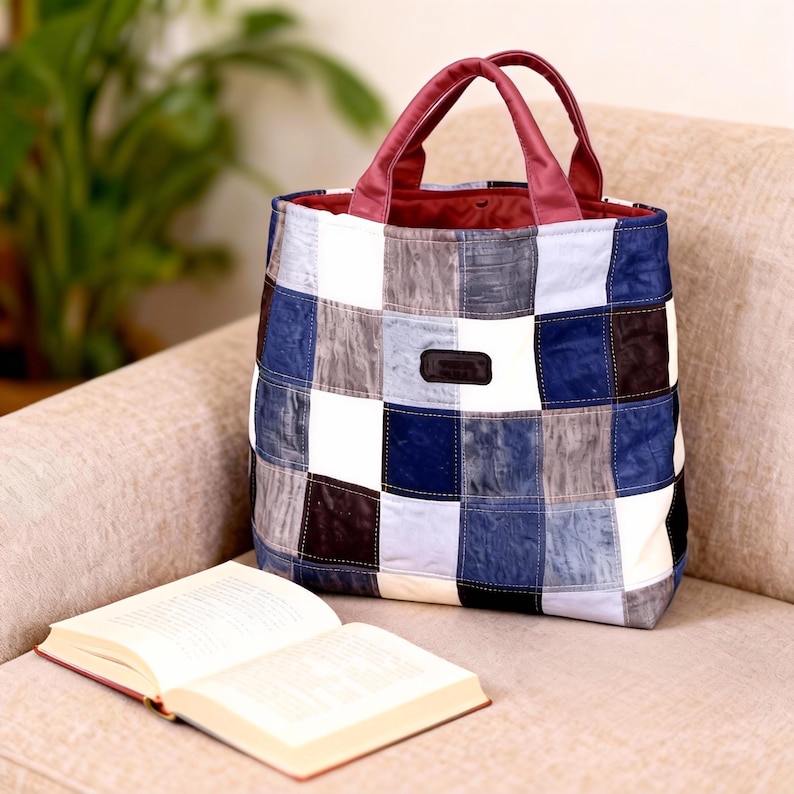 Op de afbeelding: Een patchwork tote bag met een bordeauxrode handgreep en binnenkant. De tas heeft een patroon van blauwe, witte, grijze en bruine vierkanten. Een open boek ligt naast de tas op de bank.