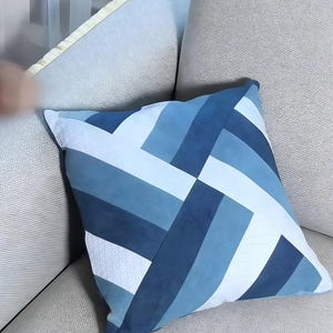 Op de afbeelding: Vierkant decoratief kussen met een geometrisch patroon in blauw- en wittinten. Het kussen heeft een patchwork-ontwerp met verschillende rechthoekige vormen, wat een moderne uitstraling cre&euml;ert. Het kussen ligt op een lichtgrijze bank.