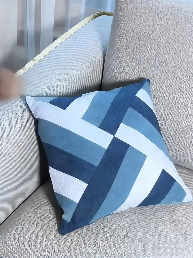 Op de afbeelding: Vierkant decoratief kussen met een geometrisch patroon in blauw- en wittinten. Het kussen heeft een patchwork-ontwerp met verschillende rechthoekige vormen, wat een moderne uitstraling cre&euml;ert. Het kussen ligt op een lichtgrijze bank.