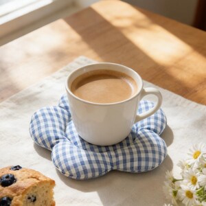 May include: A white ceramic mug filled with coffee sits on a blue and white gingham flower-shaped coaster. A slice of blueberry cake and a small bouquet of daisies are also on the table, creating a cozy, inviting scene.