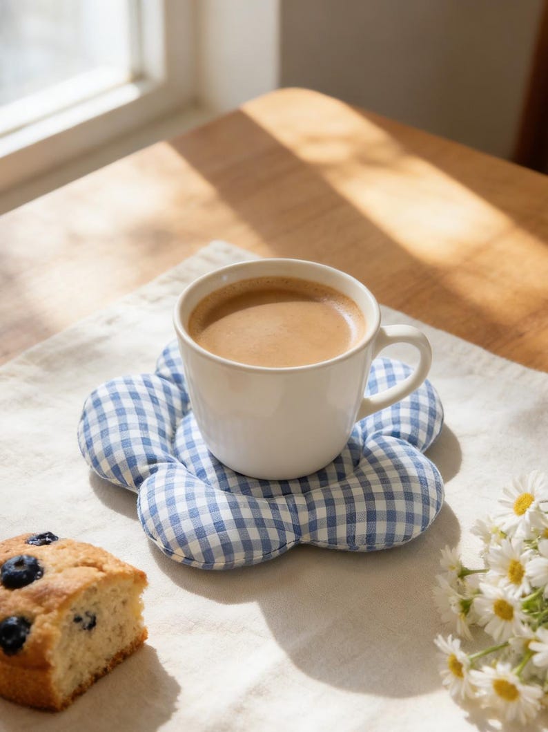 May include: A white ceramic mug filled with coffee sits on a blue and white gingham flower-shaped coaster. A slice of blueberry cake and a small bouquet of daisies are also on the table, creating a cozy, inviting scene.