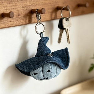 May include: A denim keychain shaped like a pumpkin wearing a dark blue denim witch hat. The pumpkin is light blue with stitched triangular eyes and a stitched mouth. It hangs from a silver-toned metal ring with a spring coil attachment.