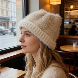 May include: A cream-colored, fluffy knit hat with a ribbed brim. The hat is worn by a person with blonde hair, seated near a window. The hat appears soft and warm, suitable for cold weather. The background shows a cafe setting.