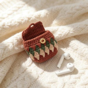 May include: A crocheted earbud case in shades of brown, green, and cream, with a gold-colored button closure. Two white earbuds rest beside the case on a cream-colored, cable-knit fabric. The case has a decorative pattern.