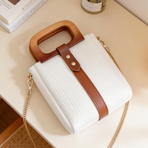 May include: White handbag with a wooden handle and a brown leather strap. The bag has a gold chain strap and a textured surface. The handle is made of light and dark brown wood.