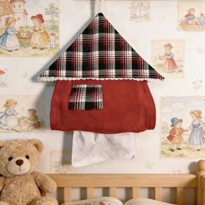 Puede incluir: Un dispensador de pañuelos de tela roja con forma de casa, con un tejado a cuadros y un pequeño cuadrado a cuadros. El borde del tejado está adornado con encaje blanco. Los pañuelos blancos son visibles en la parte inferior. Cuelga de un perchero de madera.