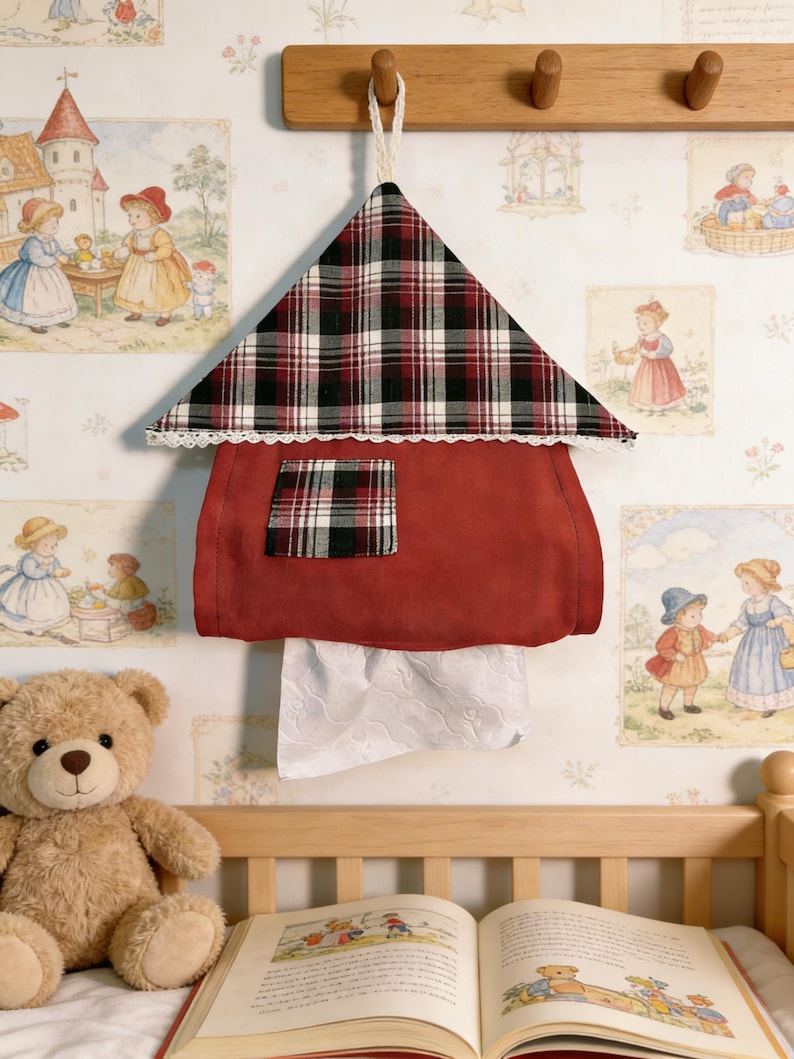 May include: A red fabric tissue holder shaped like a house, with a plaid roof and a small plaid square on the front. White lace trim edges the roof. White tissues are visible at the bottom. The holder hangs from a wooden peg rack.