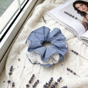 May include: A blue and white gingham scrunchie with a white eyelet lace trim. The hair accessory is set on a white knitted sweater, with sprigs of lavender and a magazine in the background.