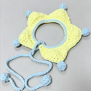 May include: A handmade, star-shaped crochet hat in yellow with a light blue trim and matching ties. The hat has light blue pom-poms at each point of the star. The hat is designed for a baby or child.