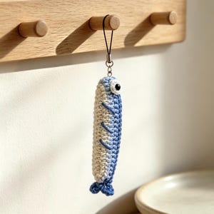 Peut inclure: Un porte-clés poisson en crochet avec un dégradé de couleurs passant du crème sur le ventre au bleu sur le dos. Le poisson possède un œil noir et blanc, des nageoires brodées en bleu et une queue bleue, fixé à une boucle en cordon noir.