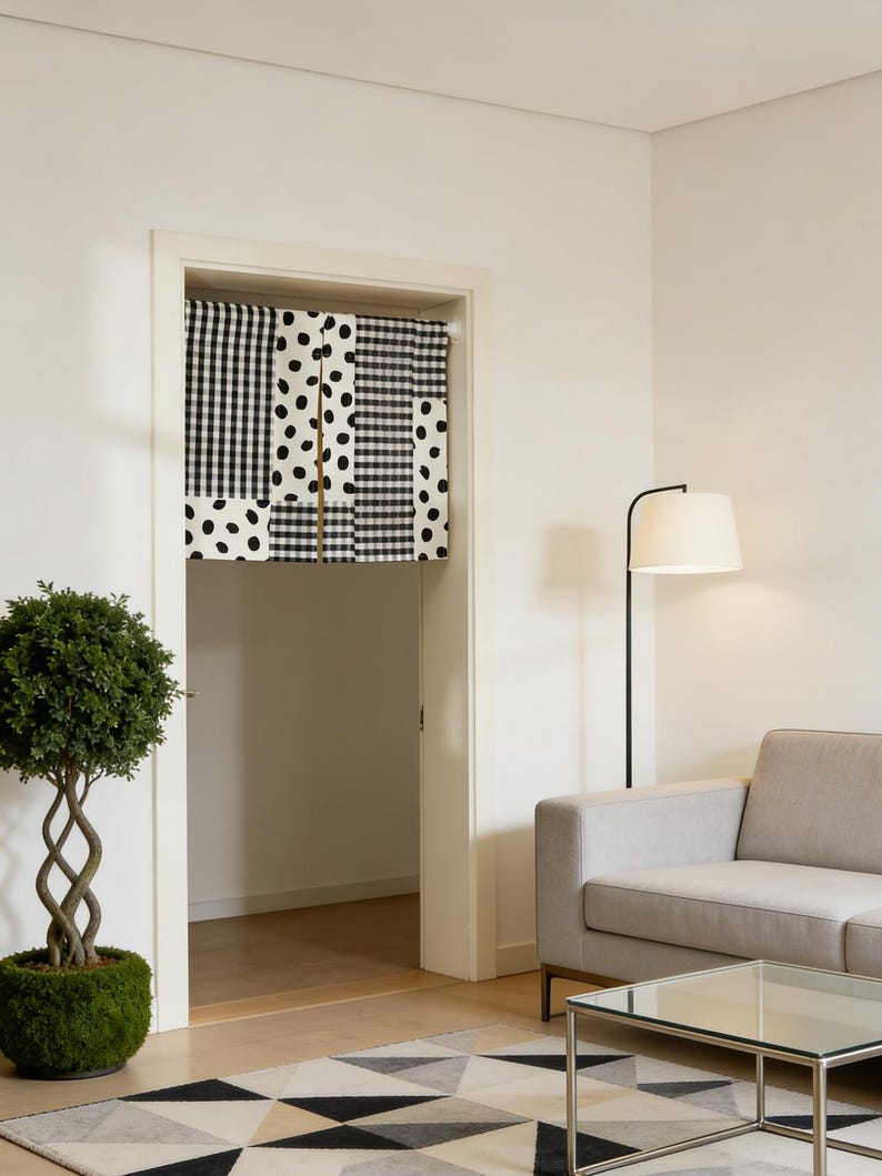 May include: A doorway curtain with a patchwork design of black and white patterns, including gingham, polka dots, and stripes. The curtain hangs in a white doorway, with a glimpse of a room beyond. The room features a sofa, a floor lamp, and a geometric rug.