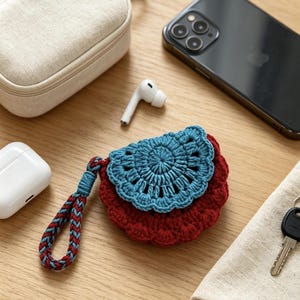 Puede incluir: Una pequeña bolsa redonda de ganchillo con una base de color rojo intenso y una solapa festoneada de color azul claro. La bolsa incluye una correa de muñeca trenzada con hilo rojo y azul, diseñada para guardar pequeños accesorios electrónicos.