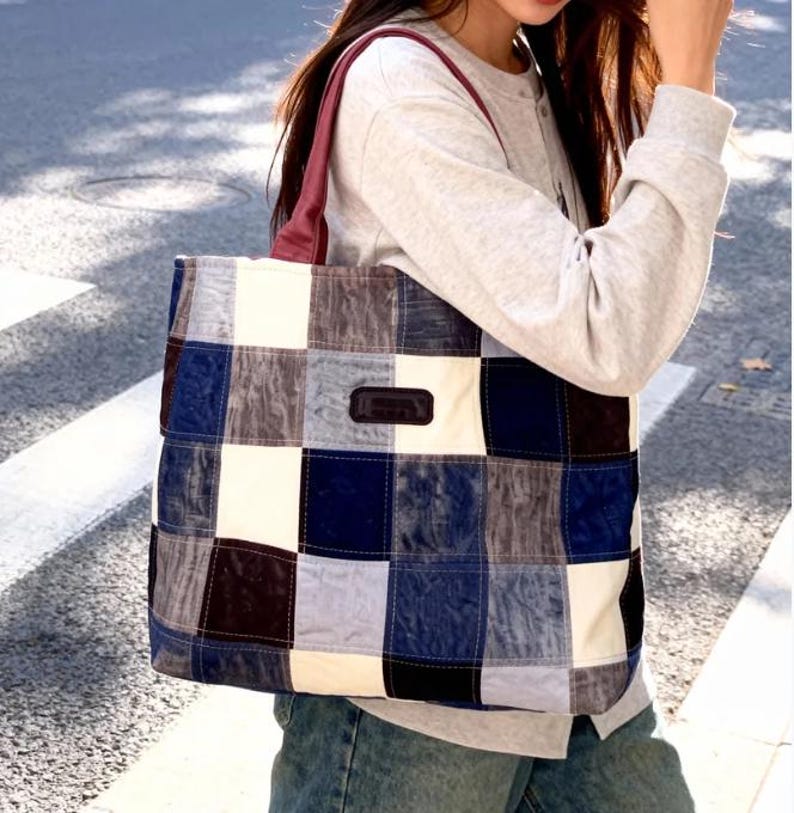 Op de afbeelding: Een patchwork tote bag met een mix van blauwe, witte en grijze vierkanten. De tas heeft een donkerbruin leren patch en bordeauxrode bandjes. De tas wordt over de schouder gedragen.