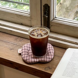 May include: A glass of iced coffee sits on a pink and white plaid coaster. The coaster is on a wooden windowsill next to an open book. The window has a view of green foliage.