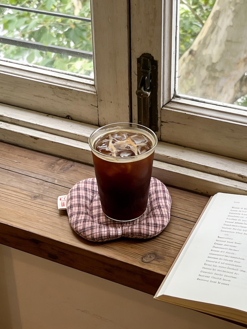 May include: A glass of iced coffee sits on a pink and white plaid coaster. The coaster is on a wooden windowsill next to an open book. The window has a view of green foliage.