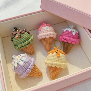Könnte beinhalten: Fünf gehäkelte Eiswaffeln in einer rosa Schachtel. Die Eiscreme-Kugeln sind in verschiedenen Farben gehalten: Grün, Rosa, Lila und Gelb, mit Streuseln und weißer Schlagsahne. Die Waffeln sind hellbraun.