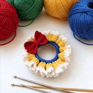 May include: A handmade crochet hair scrunchie with a red bow, yellow, white, and blue accents. Several balls of yarn in red, green, yellow, and blue are in the background, along with two crochet hooks. Craft supplies.